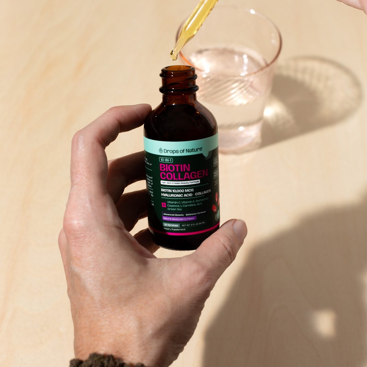 A hand holding the Biotin Collagen bottle and dropper beside a glass of water.