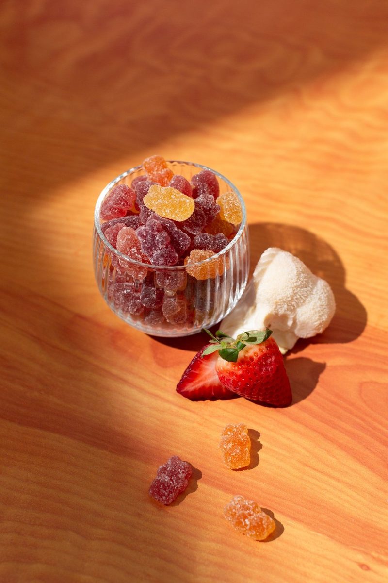 A glass of mixed fruit gummies on a wooden table with a strawberry