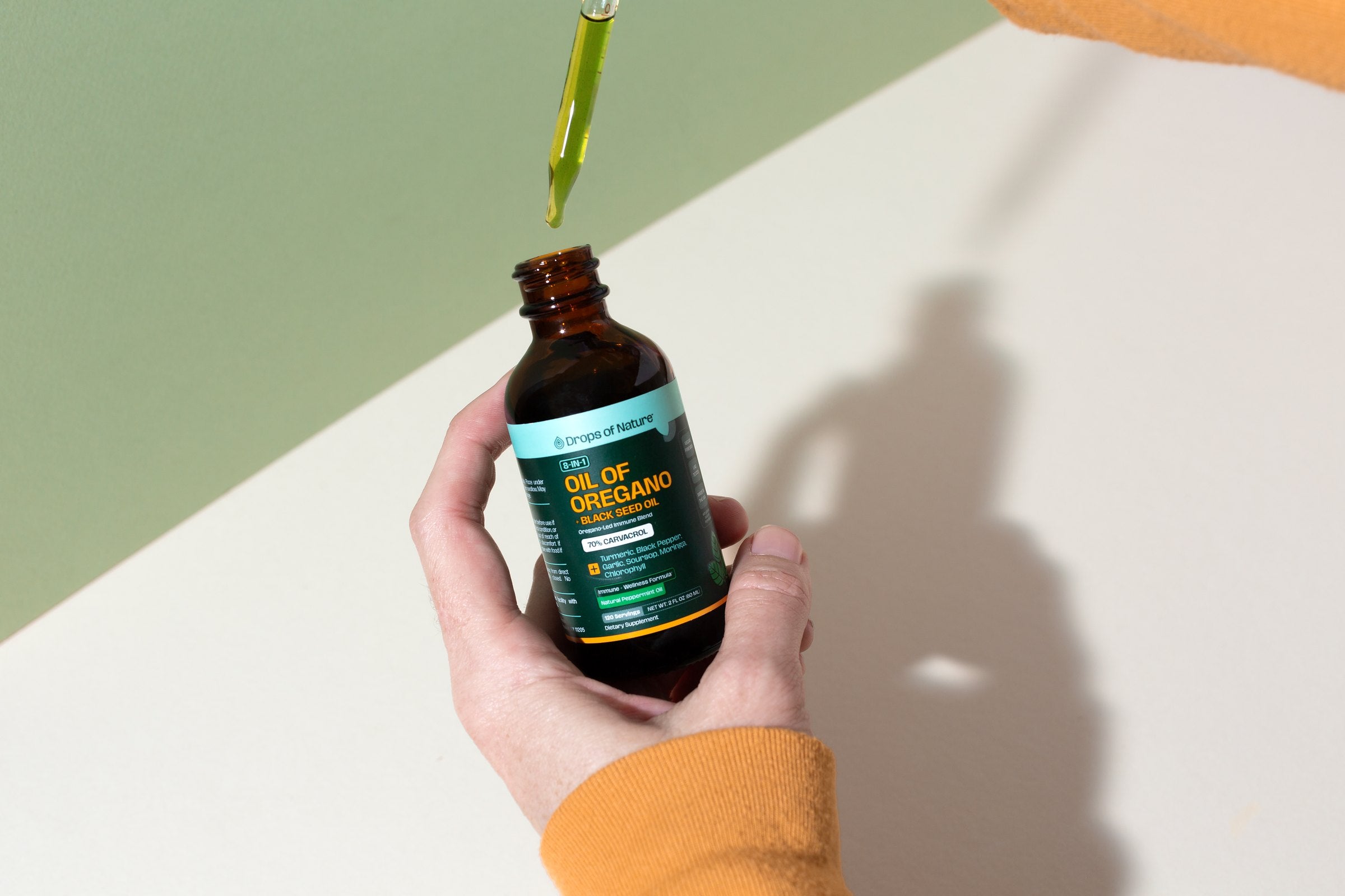 A hand holding a bottle of Oil of Oregano + Black Seed Oil as a drop falls from the dropper