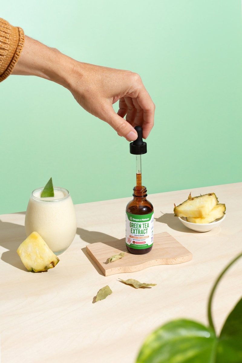 A hand lifting the dropper from a bottle of Green Tea Extract beside a pineapple smoothie