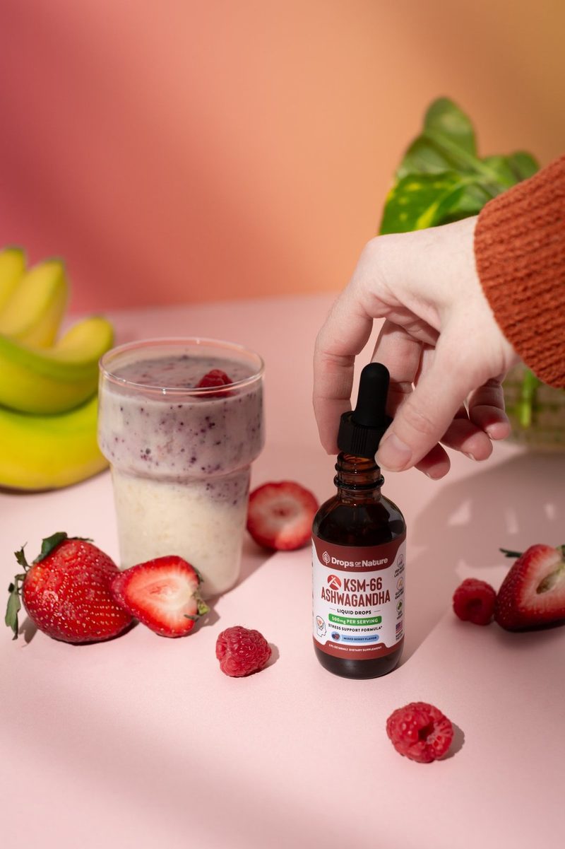 A hand reaching for KSM-66 Ashwagandha drops beside a berry smoothie