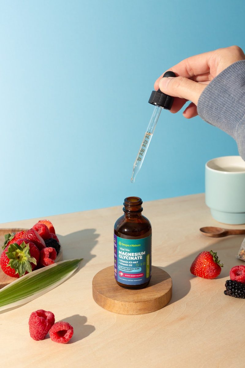 A dropper above an open bottle of Magnesium Glycinate drops on a wooden coaster, with berries