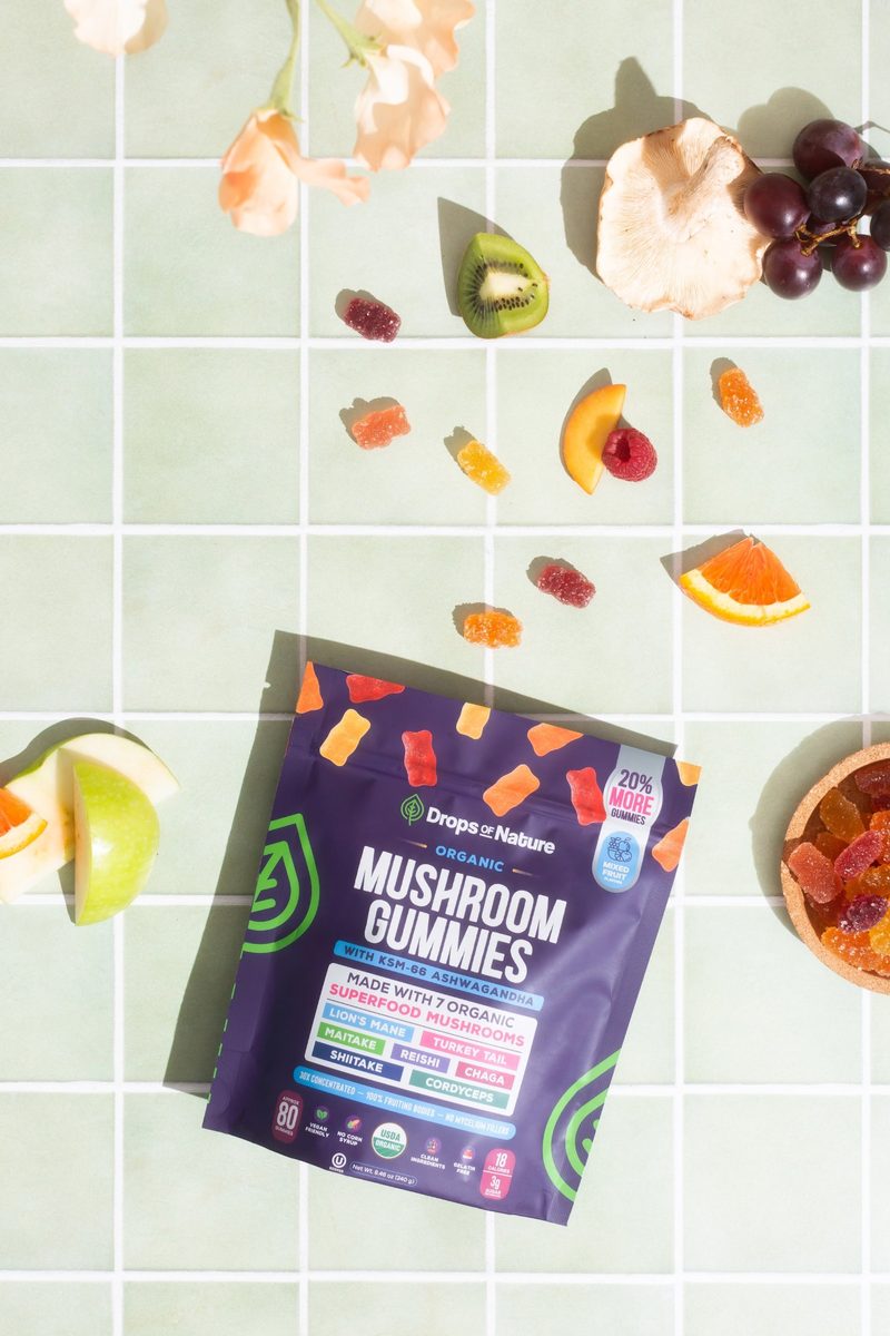 A bag of Drops of Nature Organic Mushroom Gummies on pale mint tiles, with gummies and fresh fruit scattered around it