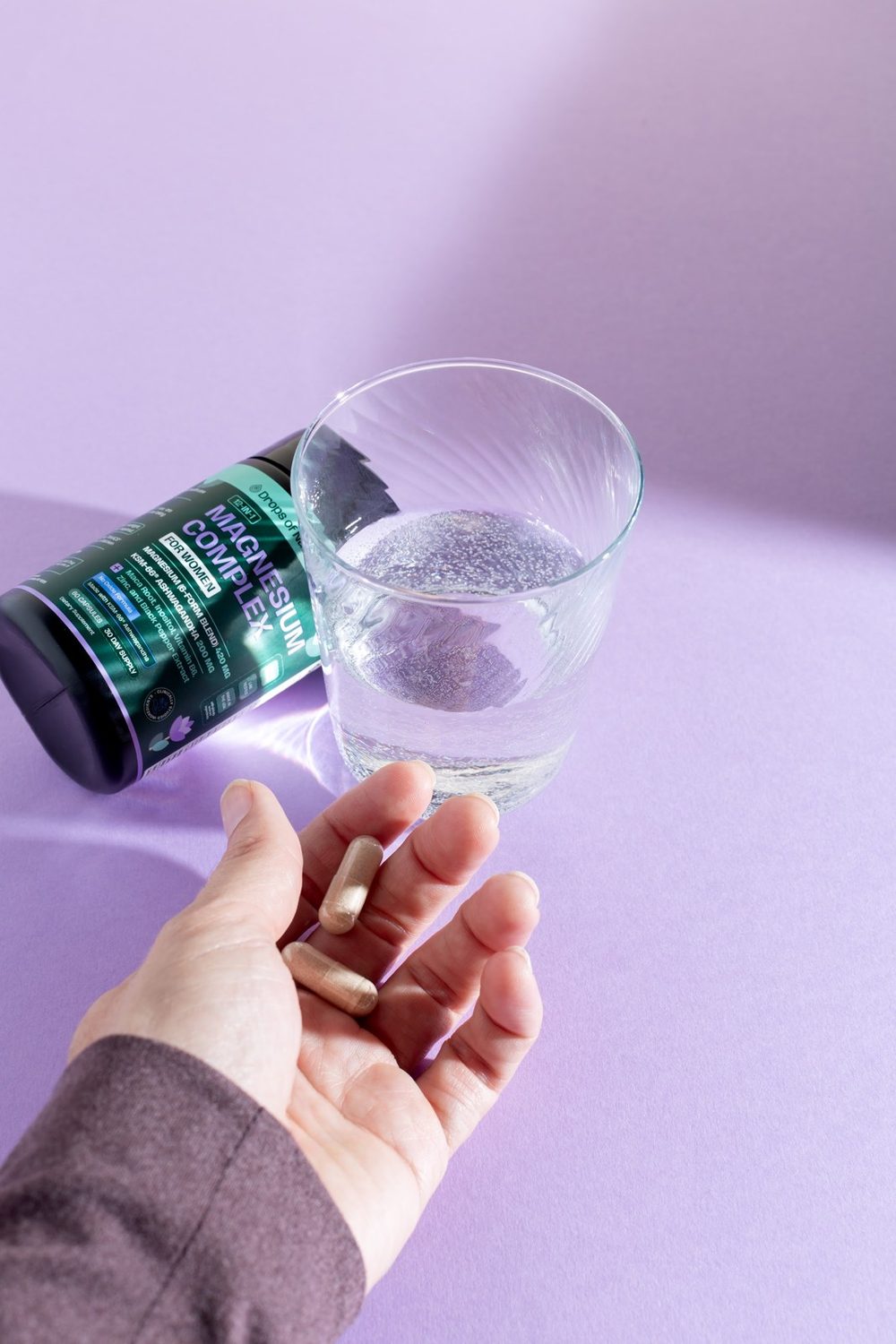 A glass of sparkling water, a bottle of 12-in-1 Magnesium Complex for Women and capsules in an open hand