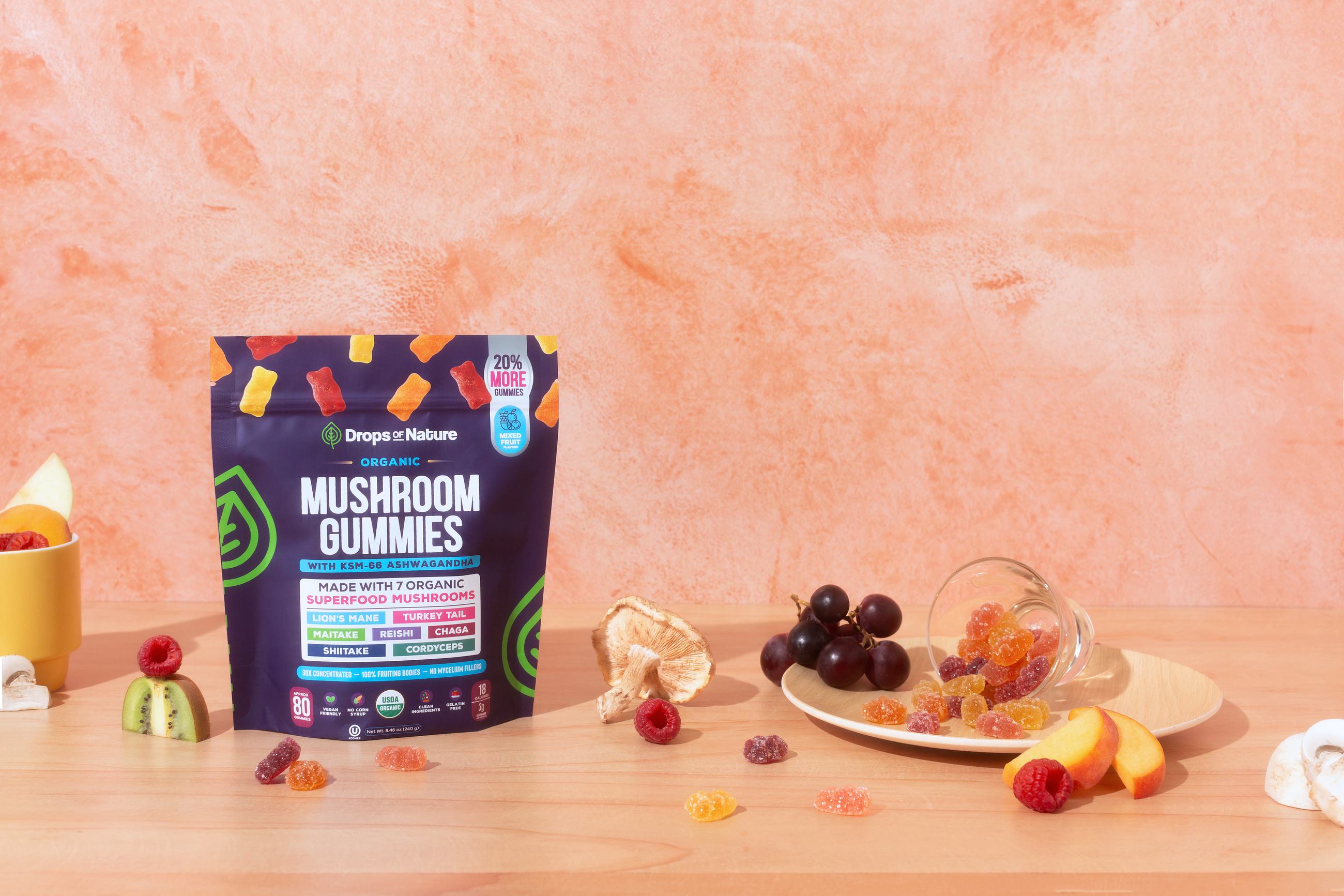 A bag of Drops of Nature Organic Mushroom Gummies on a wooden table with gummies, fruit and mushrooms