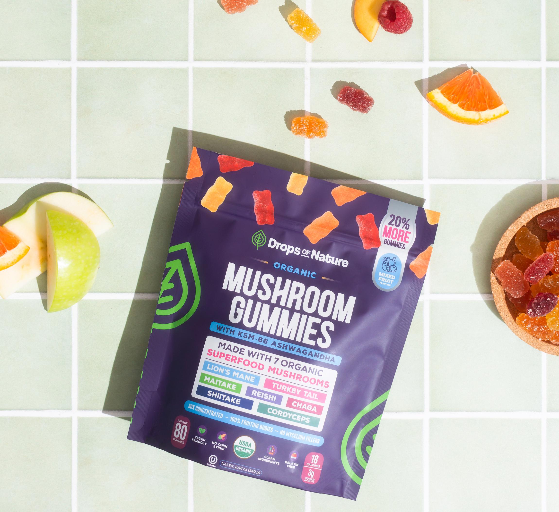 A bag of Drops of Nature Organic Mushroom Gummies on pale mint tiles, with gummies and fresh fruit scattered around it