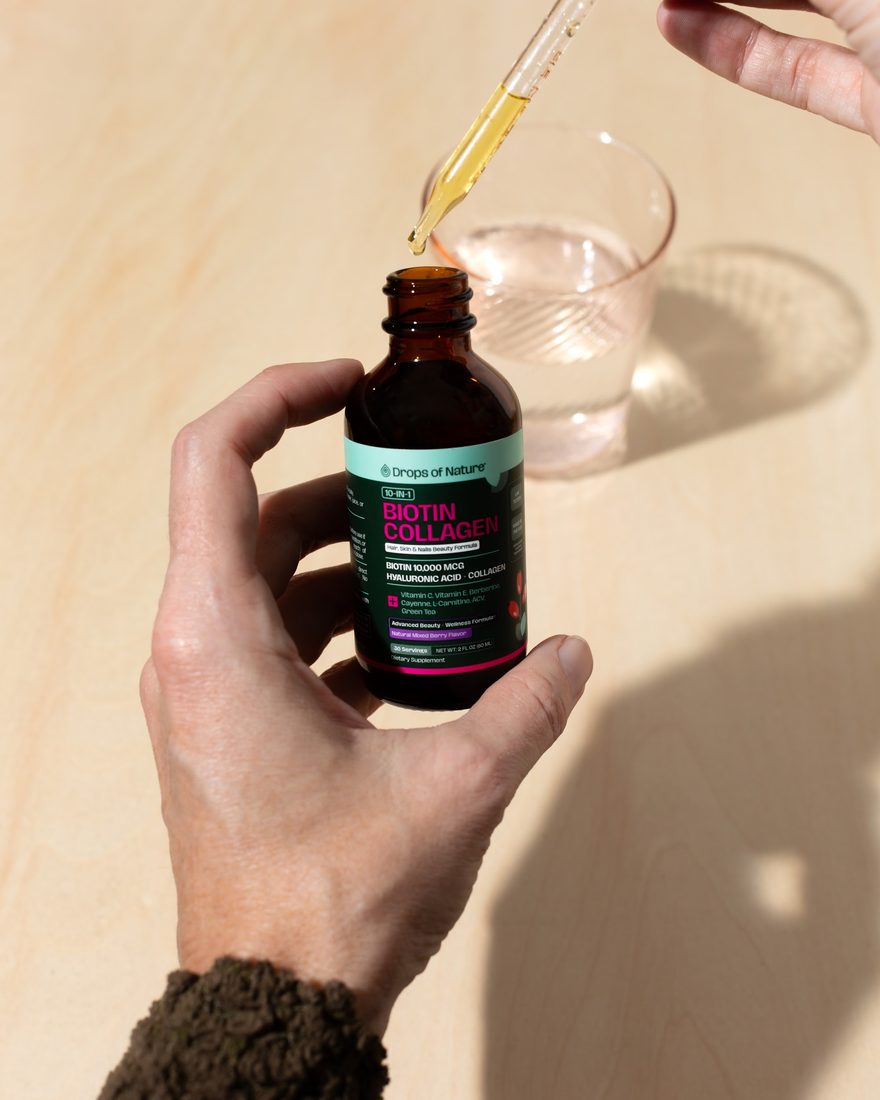 A hand holding the Biotin Collagen bottle and dropper beside a glass of water