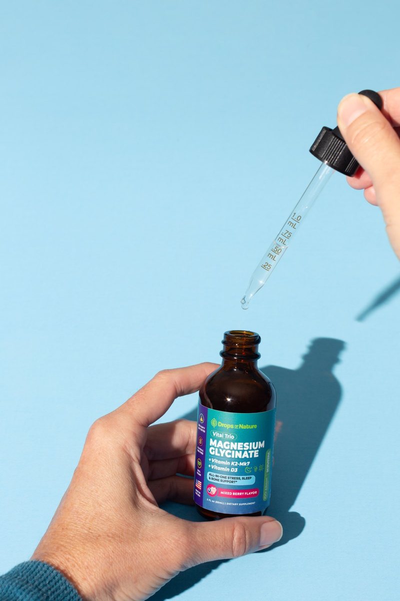 A hand holding a bottle of Magnesium Glycinate + D3 & K2 drops.