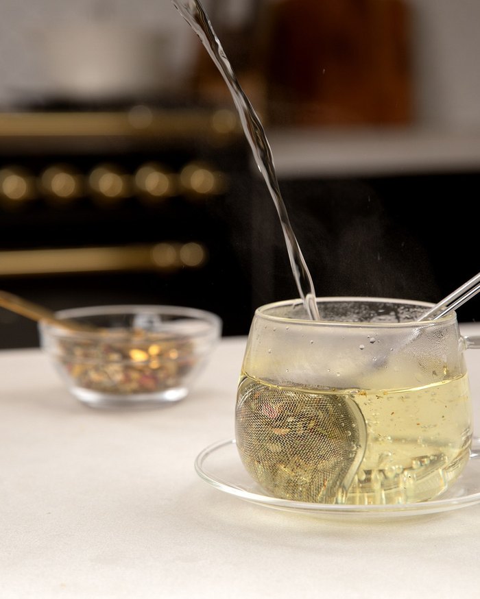 Loose-leaf tea being poured into a glass cup, with a Pella loose-leaf tea pouch behind